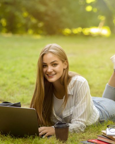 Online Education And Distance Learning. Beautiful Ten Girl Studying With Laptop Outdoors, Lying On Lawn In Park, Smiling At Camera, Copy Space